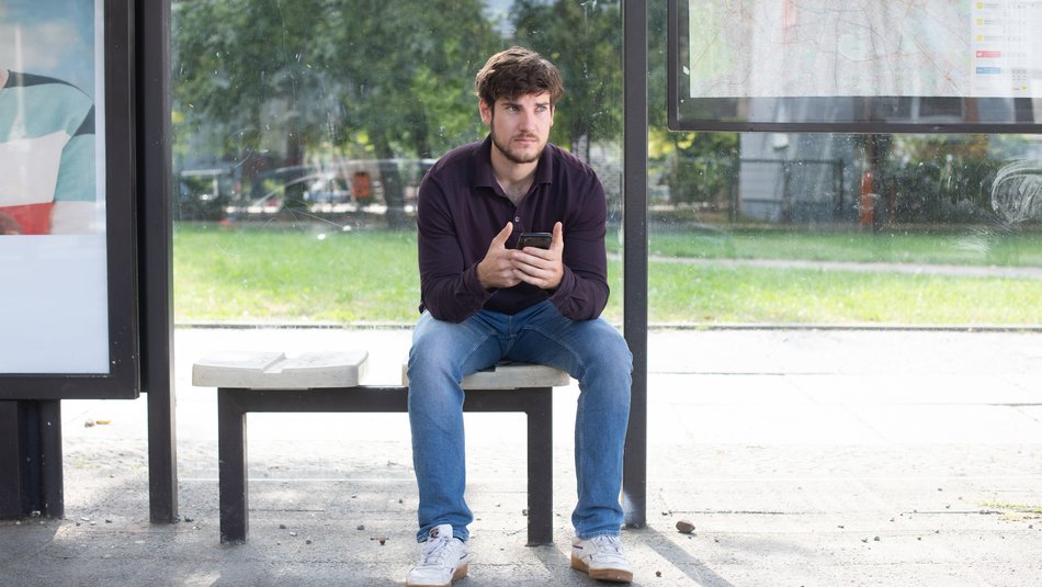 Ein junger Mann sitzt an einer Bushaltestelle, er hat ein Smartphone in der Hand
