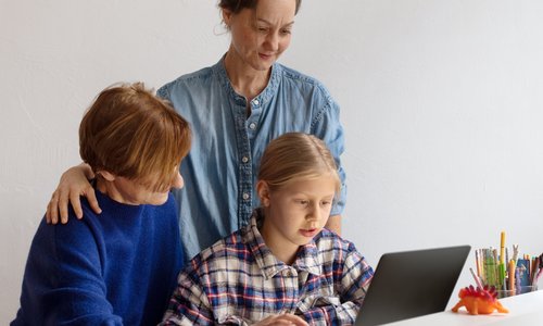 Zwei Mütter und ein Kind sitzen bzw. stehen vor einem Laptop und schauen auf den Bildschirm