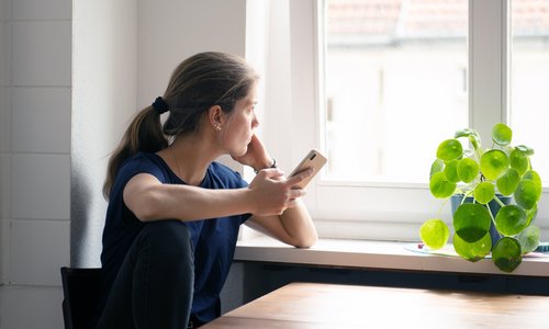 Eine junge Frau schaut aus dem Fenster, in der Hand hält sie ein Telefon