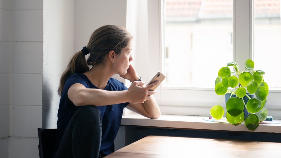 Eine junge Frau schaut aus dem Fenster, in der Hand hält sie ein Telefon