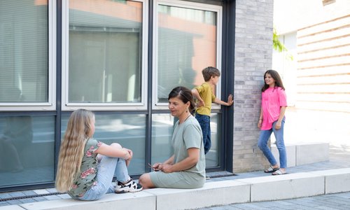Vor einem Gebäude stehen zwei Kinder, ein weiteres Kind sitzt auf einem Mauervorsprung und unterhält sich mit einer erwachsenen Person.