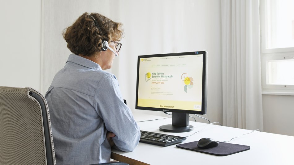 Frau sitzt vor einem Computer am Schreibtisch. Auf dem Desktop sieht man die Website www.hilfe-telefon-missbrauch.de 